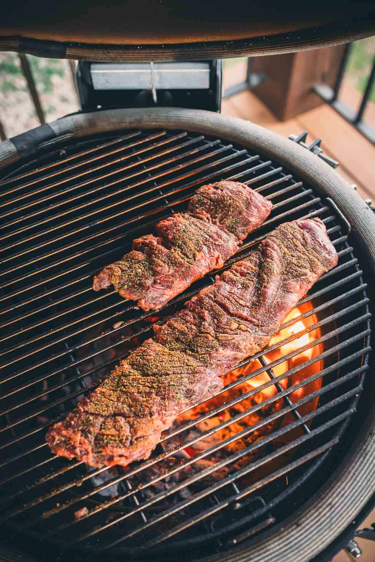 Two seasoned skirt steaks cooking on a grill with visible flames beneath.