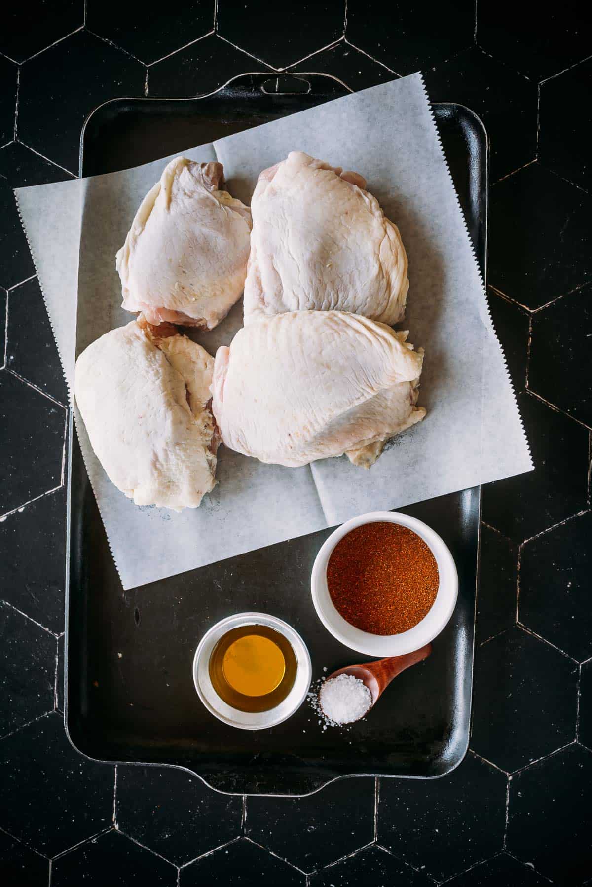 A black tray holds four raw chicken thighs on parchment paper, accompanied by bowls of oil, spice mix, and a wooden spoon with salt.