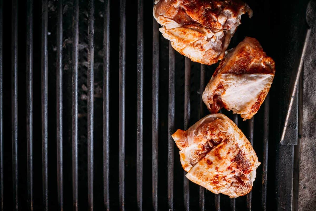 Three pieces of grilled chicken with browned, slightly crispy skin are arranged on a black grill with wide, parallel bars.