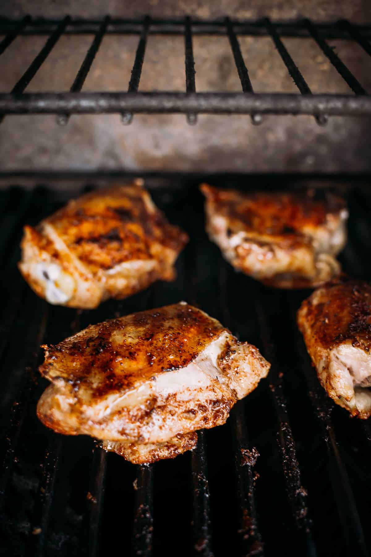 Four seasoned chicken thighs are being grilled on a barbeque grill. The pieces are golden brown and show visible grill marks.