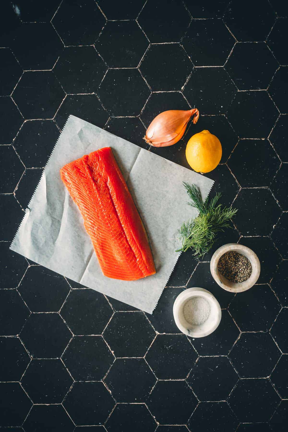 A fillet of raw salmon on parchment paper, accompanied by a shallot, a lemon, dill, a bowl of salt, and a bowl of pepper, all placed on a black hexagonal-tiled surface.