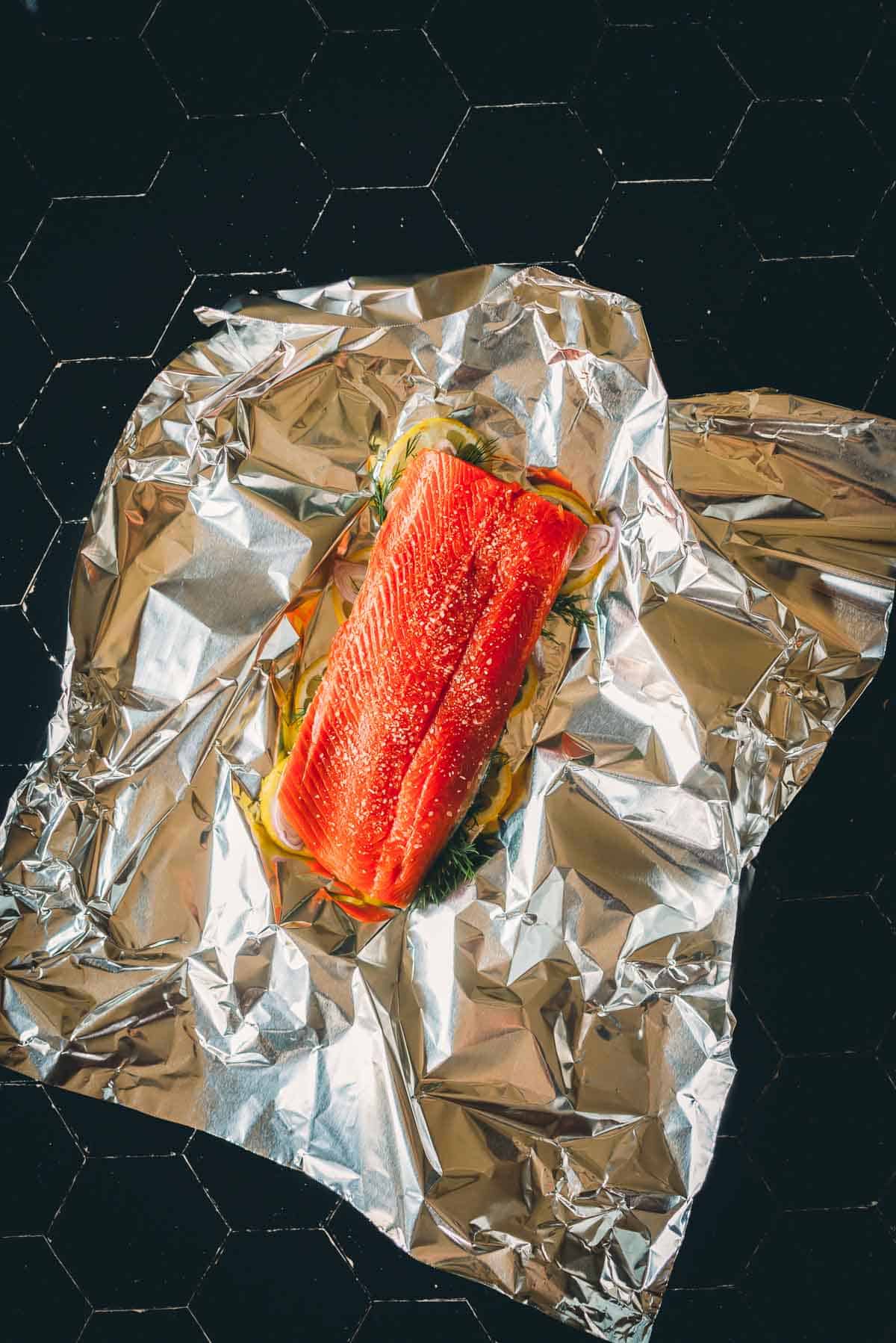 A piece of raw salmon fillet is placed on a sheet of aluminum foil with seasonings and herbs, ready to be cooked.