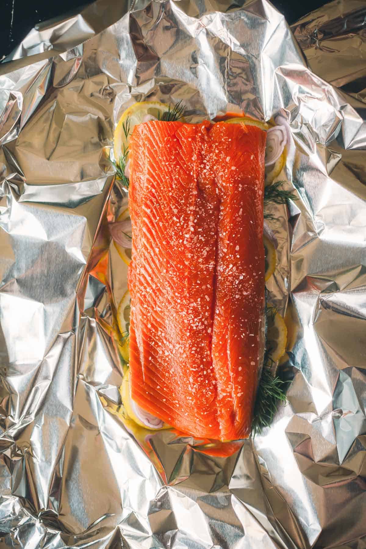 A raw salmon fillet on foil, seasoned with salt and placed on a bed of lemon slices and herbs.