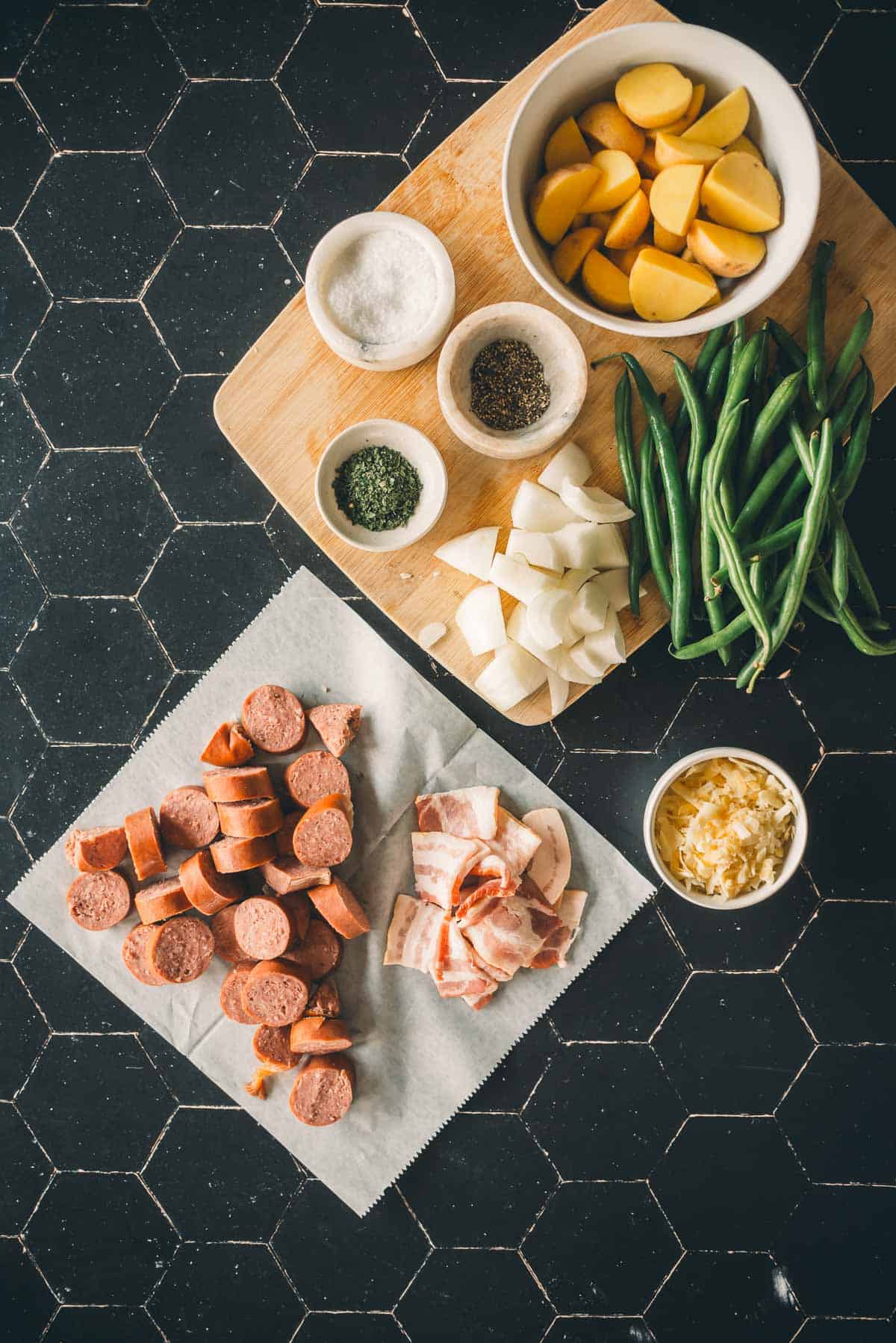 Ingredients on a hexagonal-tiled counter include sliced sausage, bacon pieces, chopped potatoes, green beans, diced onions, shredded cheese, salt, pepper, and herbs on a cutting board.