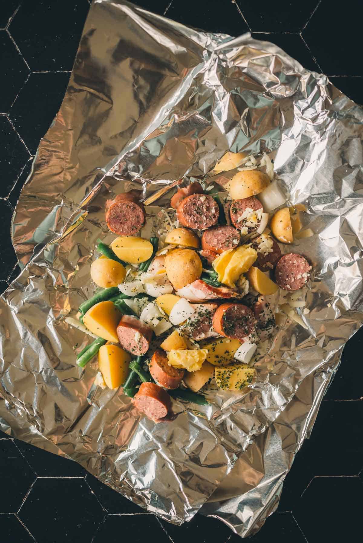 A foil packet contains cooked sausage slices, diced potatoes, green beans, and seasonings on a black hexagon-tiled surface.