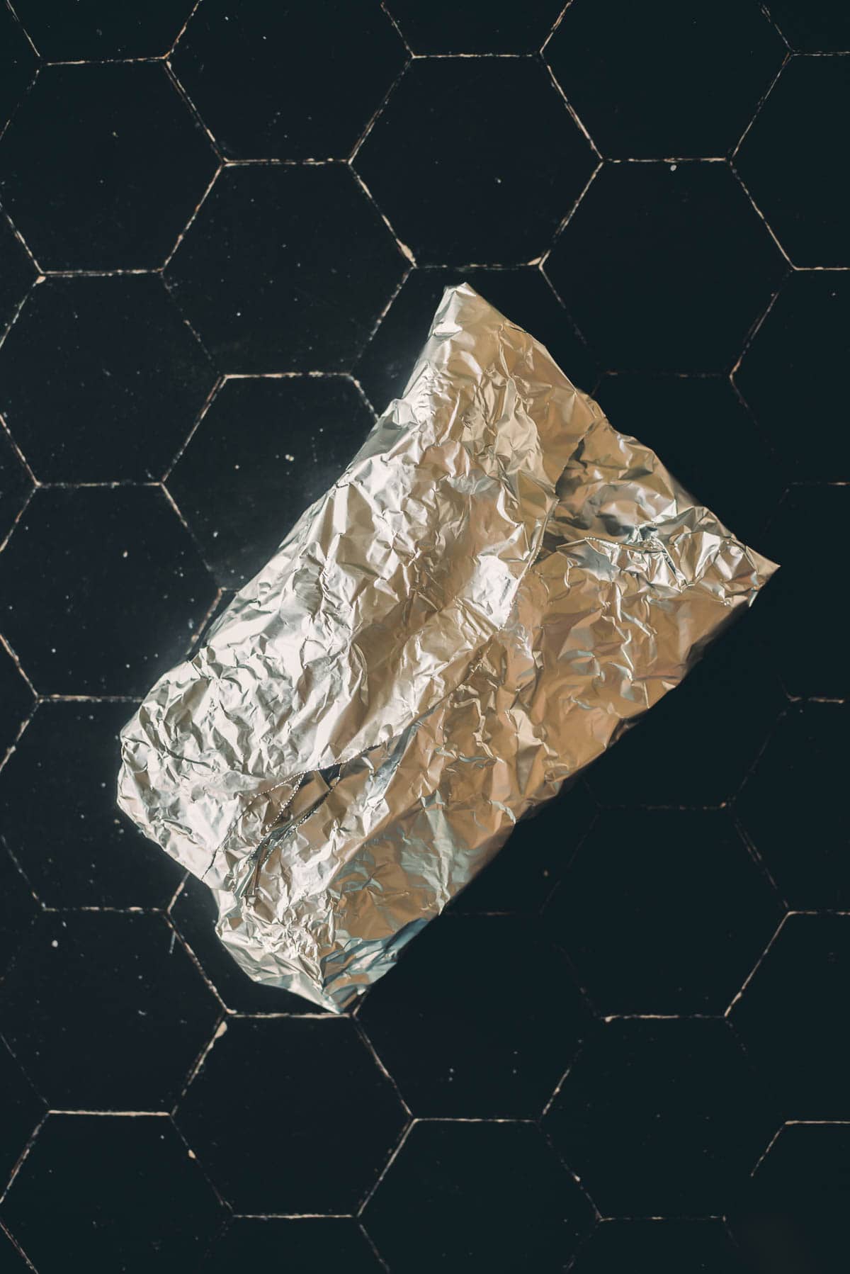 A crumpled piece of aluminum foil lies on a black hexagonal-tiled surface.