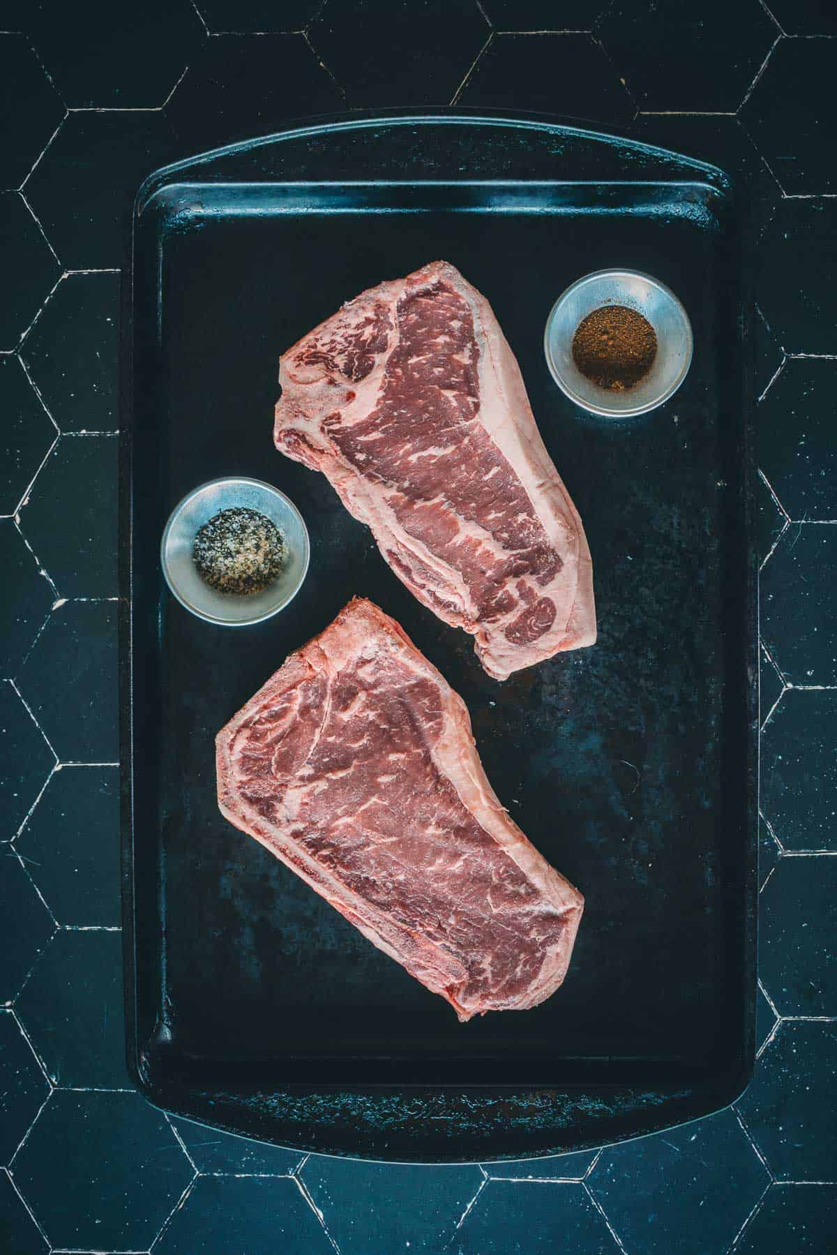 Two raw strip steaks on a black baking tray with a small bowl of pepper and a small bowl of seasoning, placed on a tiled surface.