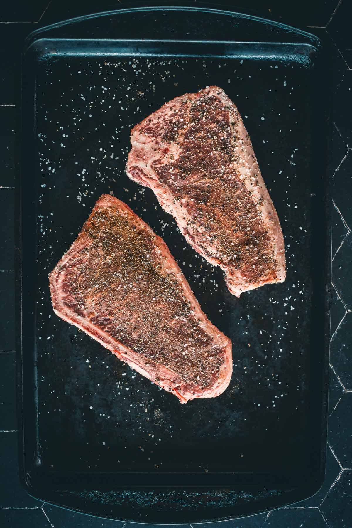 Two raw, seasoned steaks placed on a black baking tray, ready for cooking.