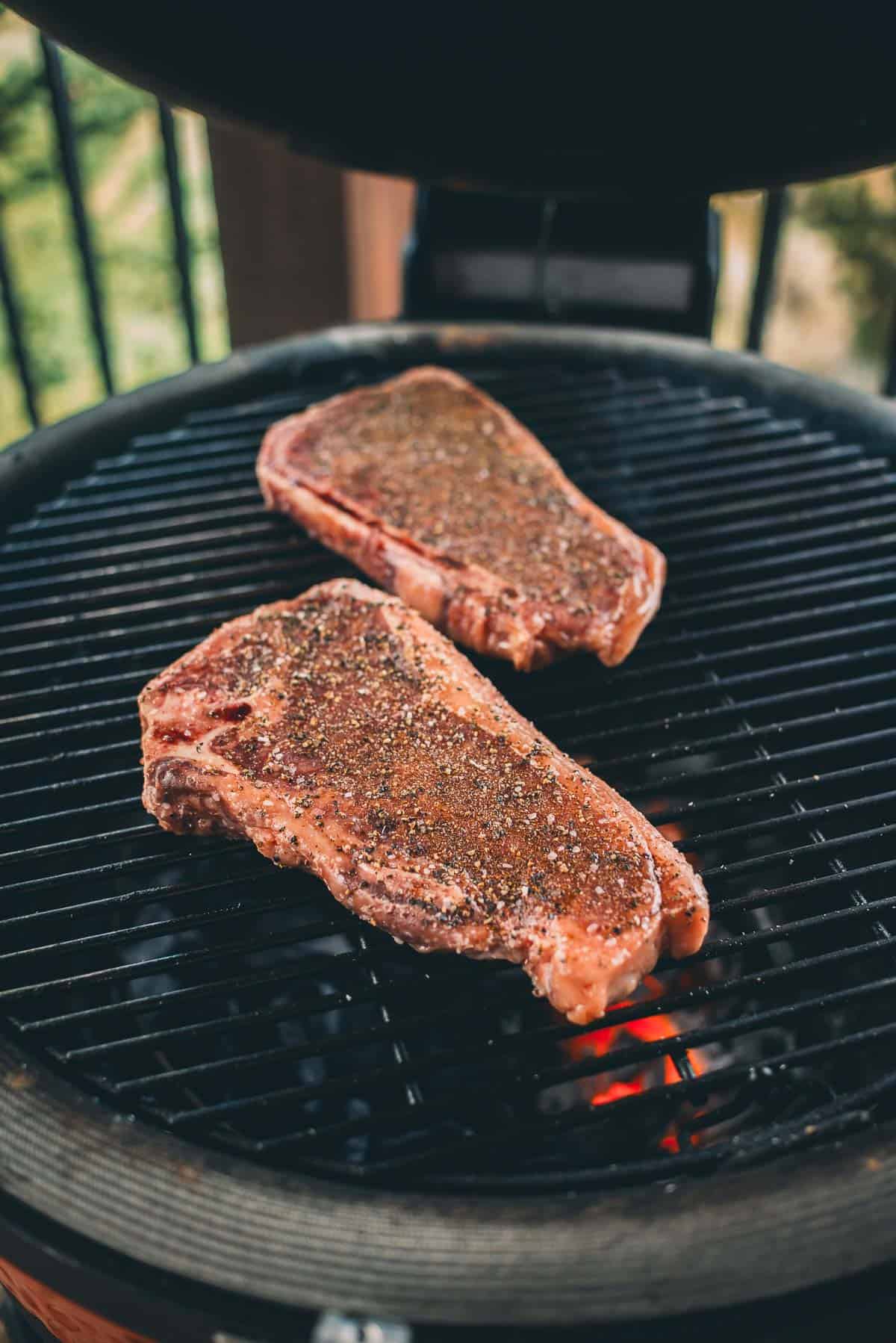 Two seasoned strip steaks cooking on a grill with visible flames underneath.