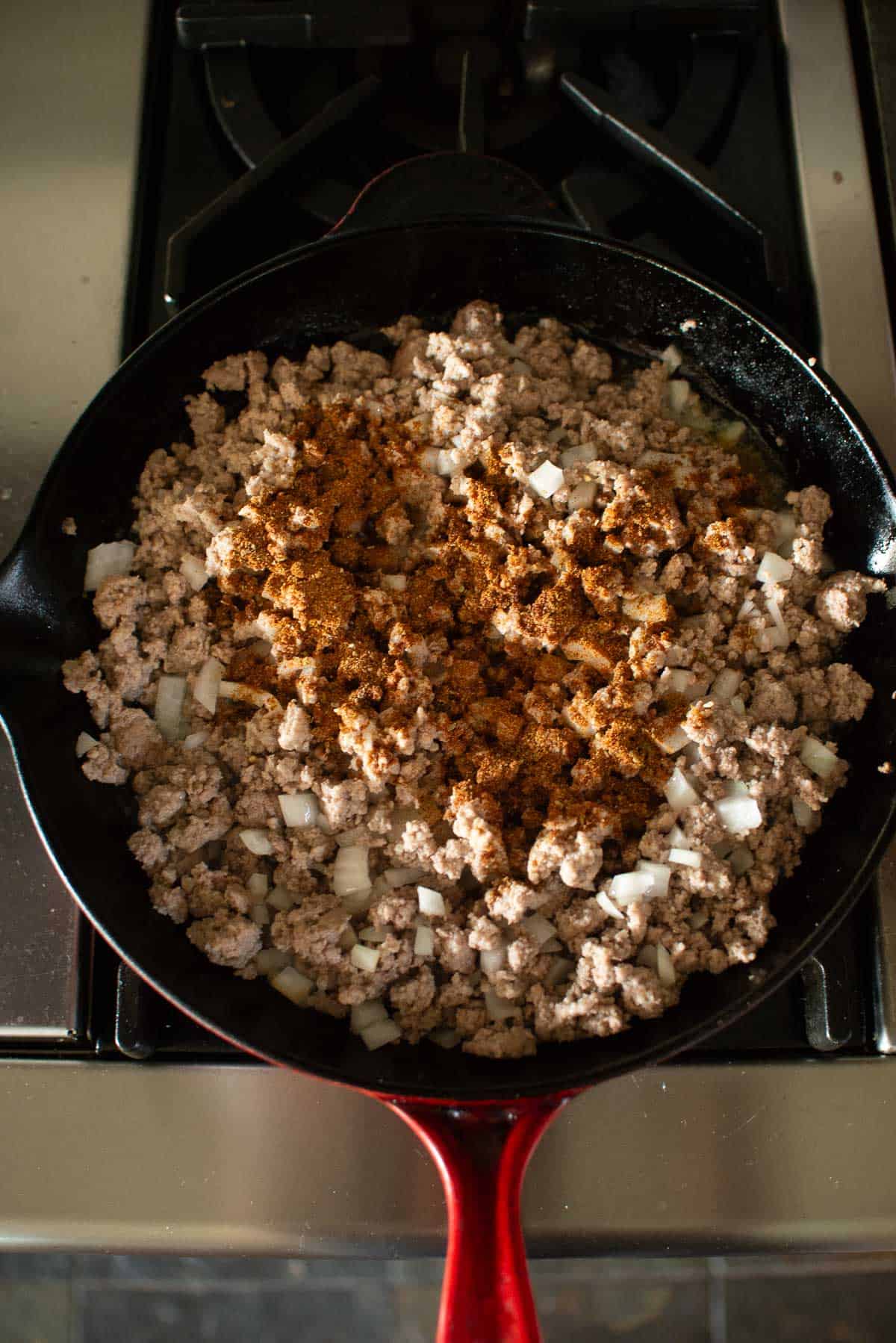 A cast iron skillet on a stove containing ground meat, diced onions, and a heap of brown seasoning.