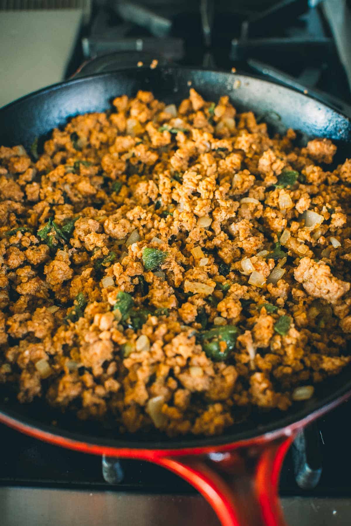 A close-up of crumbled, seasoned ground meat being cooked in a red skillet on a stovetop.