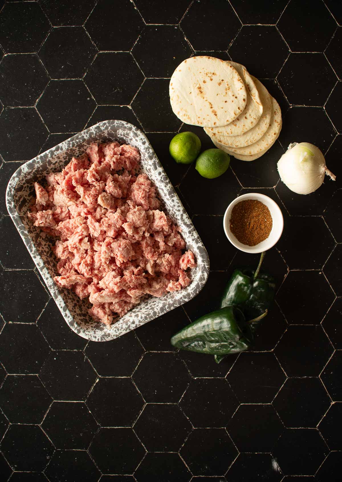 A tray of ground meat, a stack of tortillas, two limes, an onion, a small bowl of spices, and a poblano pepper are arranged on a black hexagonal-tiled surface.