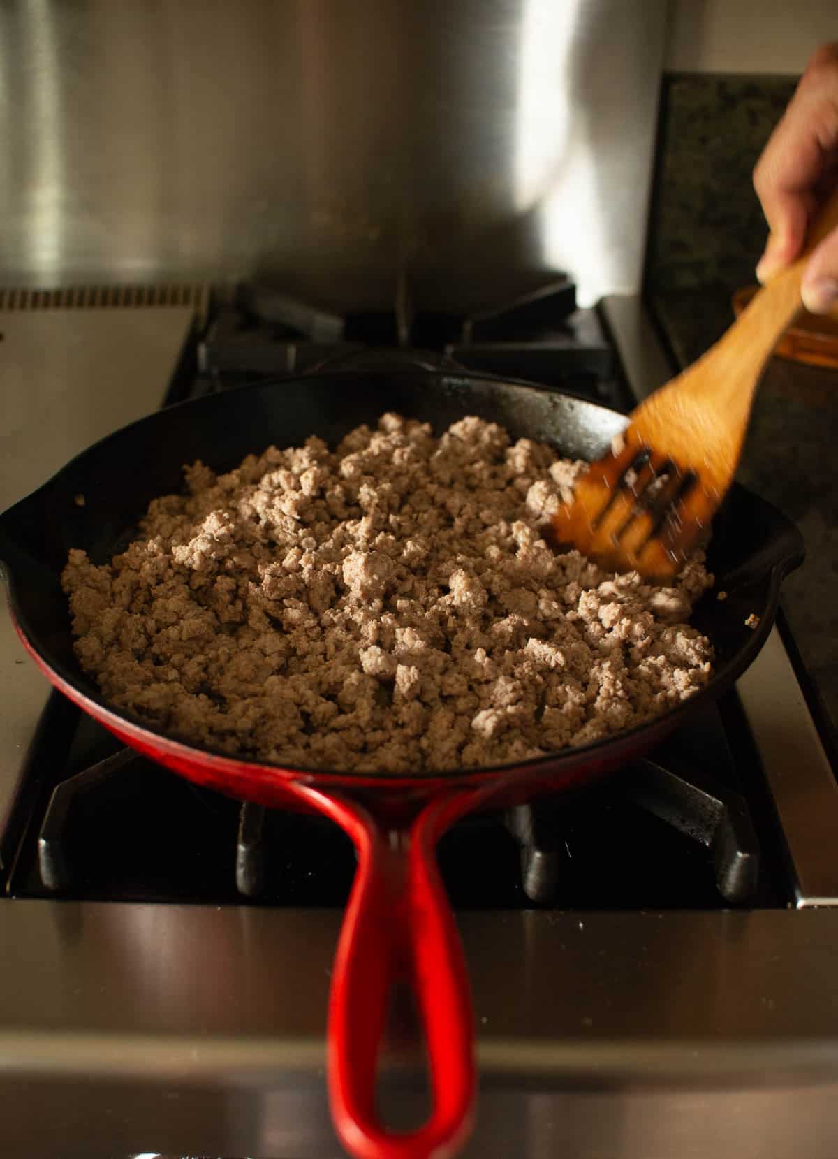 A person is stirring ground meat in a red cast iron skillet on a stove using a wooden spatula.