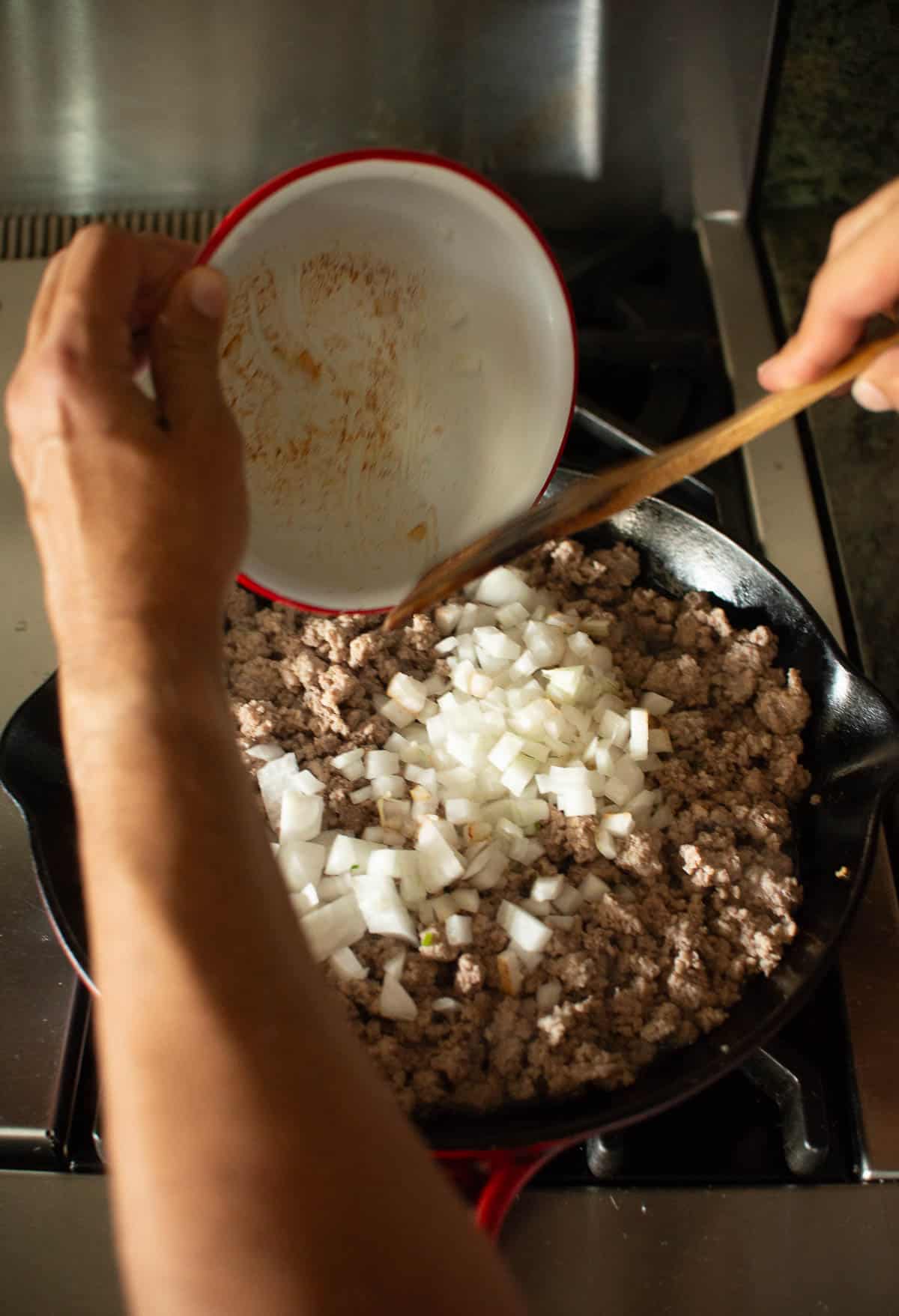 Person adding chopped onions from a red bowl into a skillet filled with browned ground meat on a stovetop.