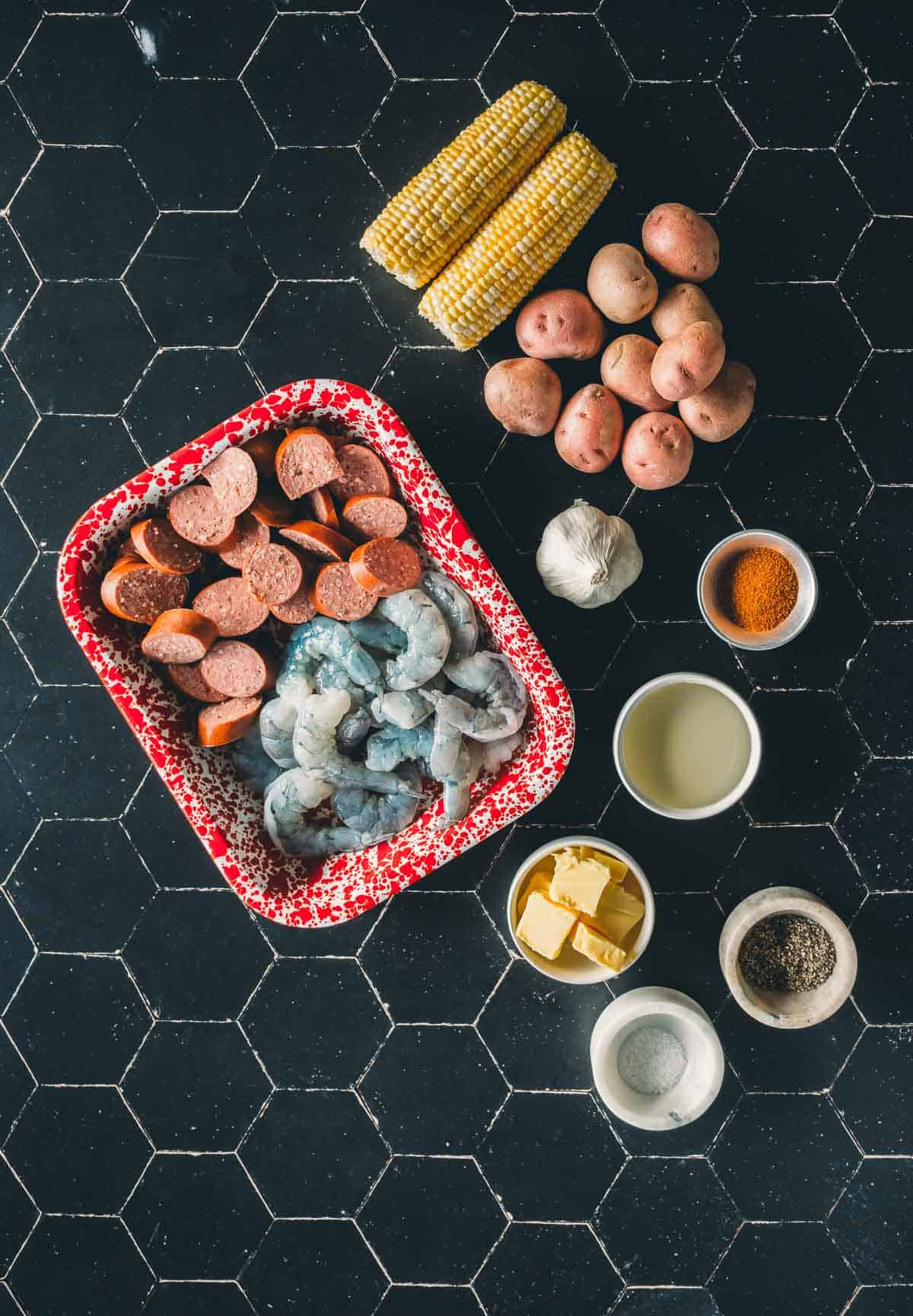 Ingredients for a shrimp boil: uncooked shrimp, sliced sausage, corn on the cob, potatoes, garlic, seasoning, butter, oil, and spices arranged on a black hexagonal tile surface.