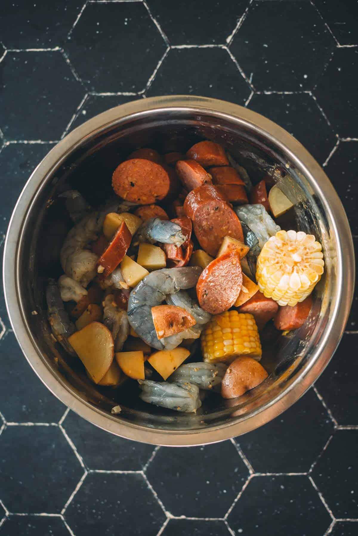 A metal bowl containing a mix of uncooked shrimp, sausage slices, potato chunks, and corn on the cob, placed on a black hexagonal-tiled surface.