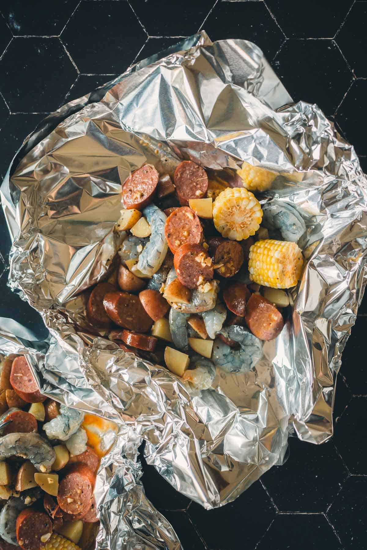 Two foil packets containing a mix of cooked sausage, shrimp, corn, and diced potatoes on a dark hexagonal tile surface.