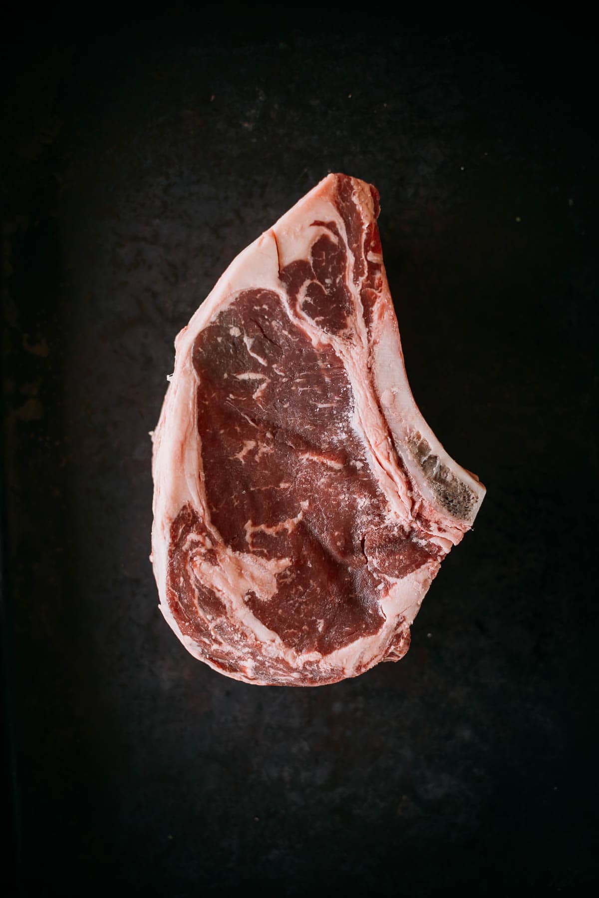 A raw cut of ribeye steak with a visible bone on a dark surface.