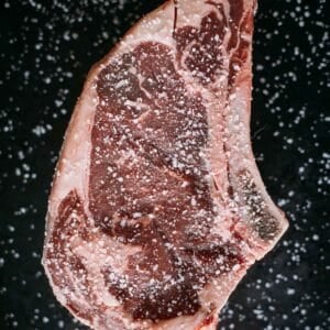 Raw ribeye generously seasoned with salt on a dark surface.