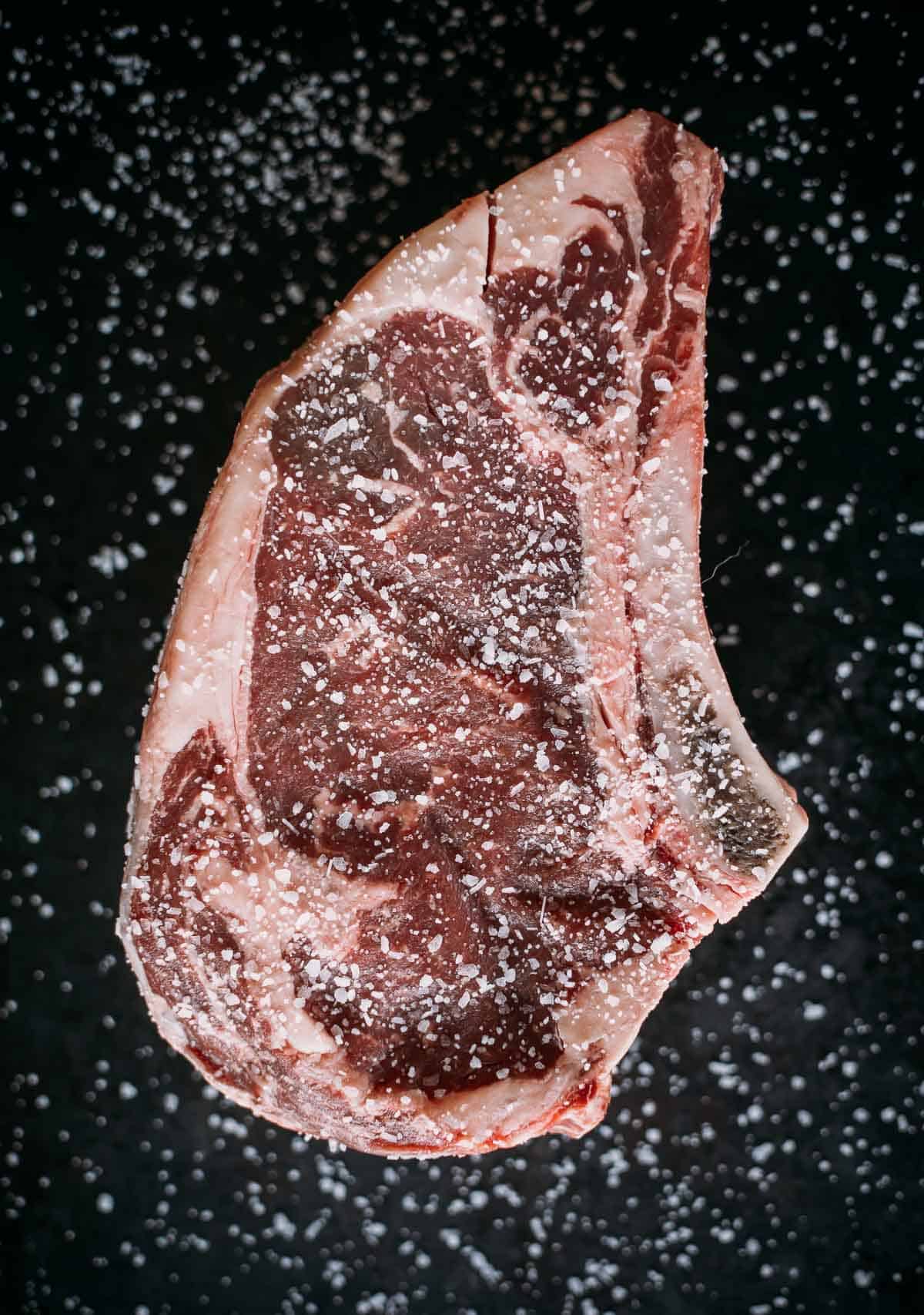 Raw ribeye generously seasoned with salt on a dark surface.