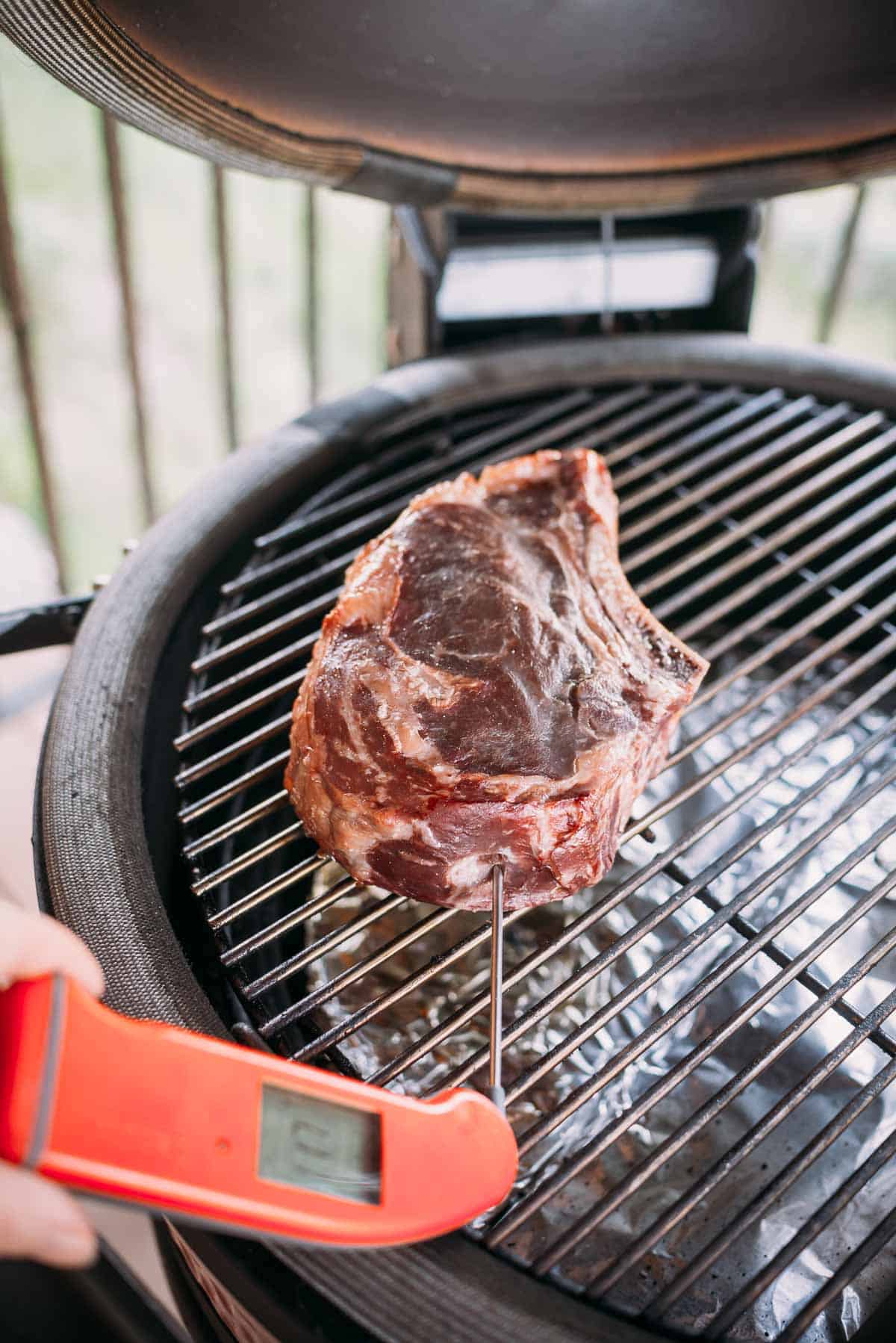 A hand holds a food thermometer inserted into a large steak cooking on a grill with the lid open.