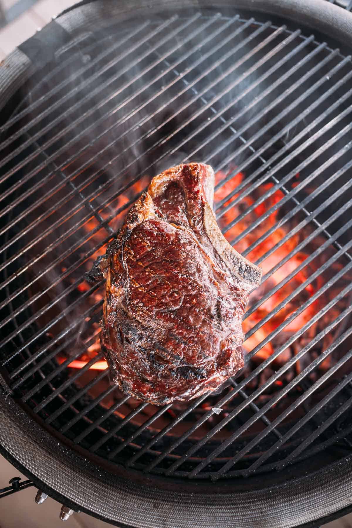 A large steak grilling on an open flame barbecue with visible char marks.