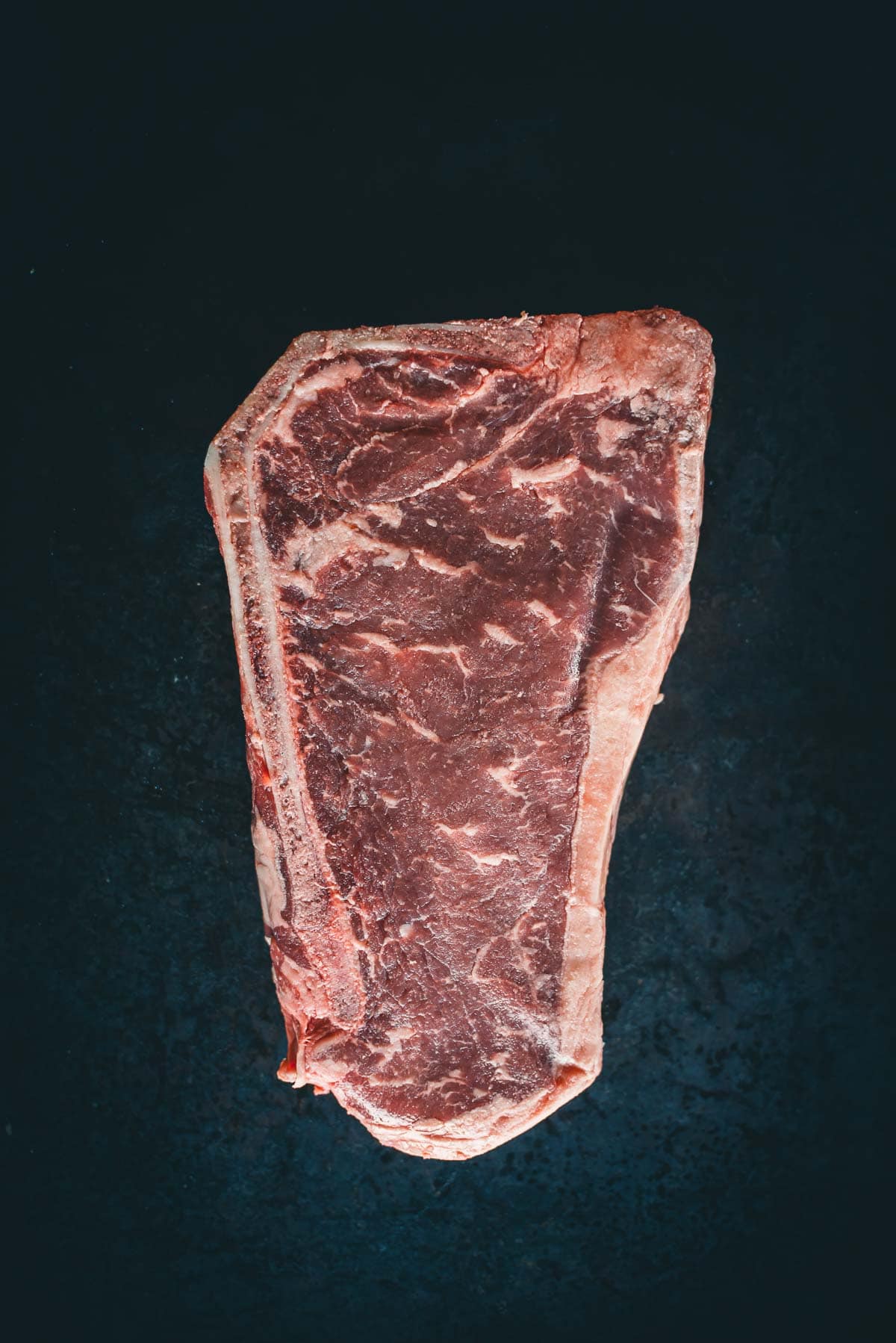 A raw Kansas City strip steak with visible marbling and a layer of fat around the edges, against a dark background.