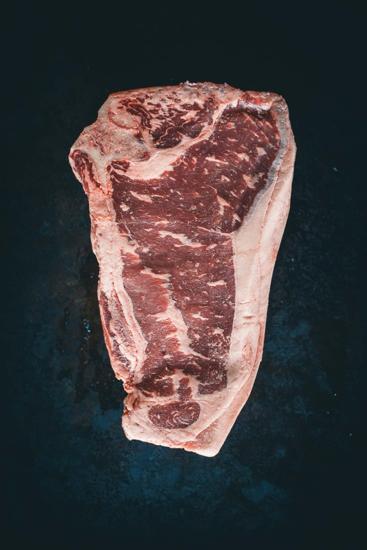 A raw, marbled Kansas city strip steak is shown against a dark background.