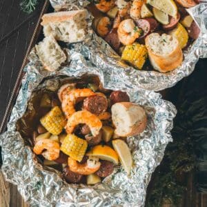 Two foil packets containing shrimp, sausage, corn, and potatoes garnished with lemon wedges and served with pieces of bread.