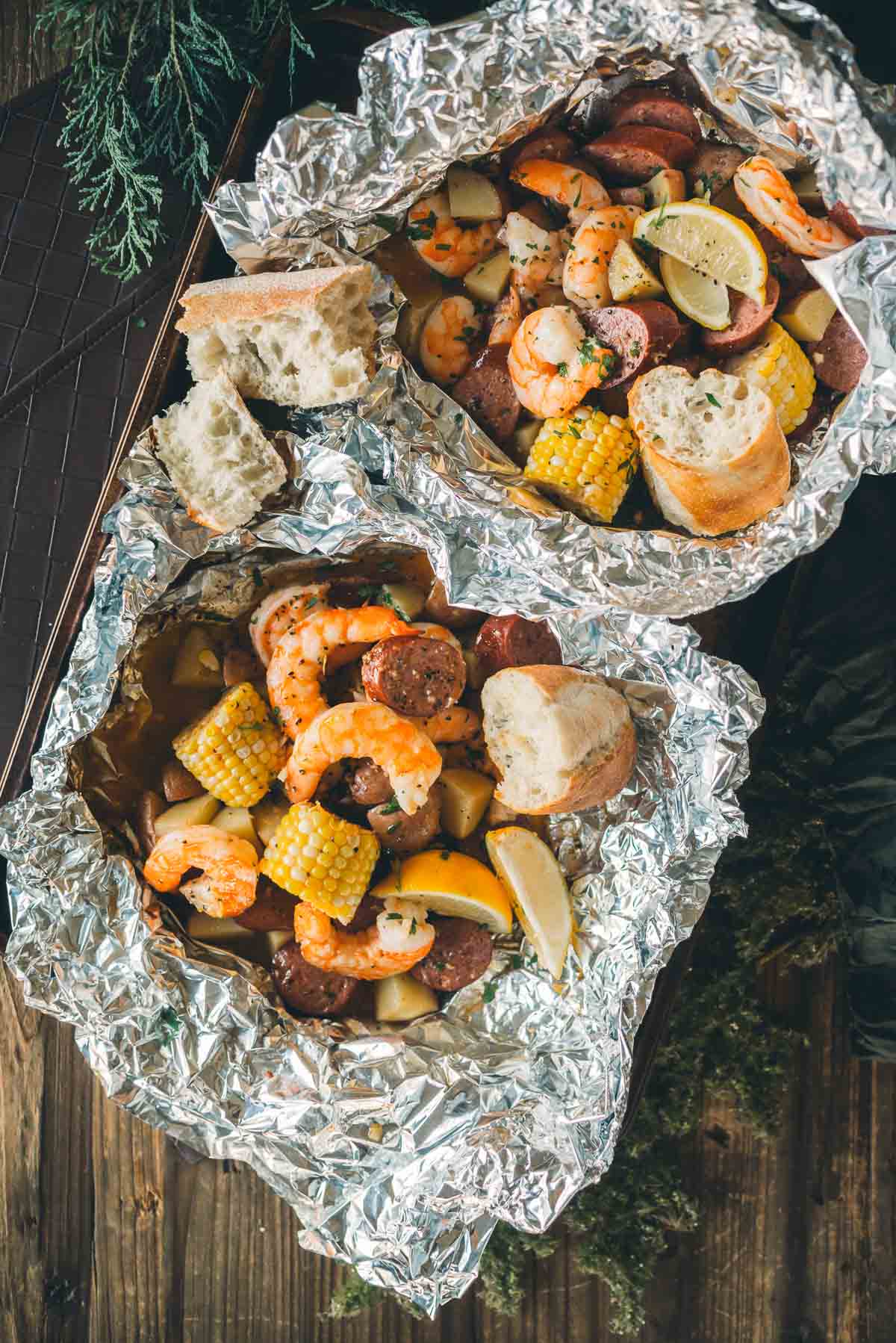 Two foil packets containing shrimp, sausage, corn, and potatoes garnished with lemon wedges and served with pieces of bread.