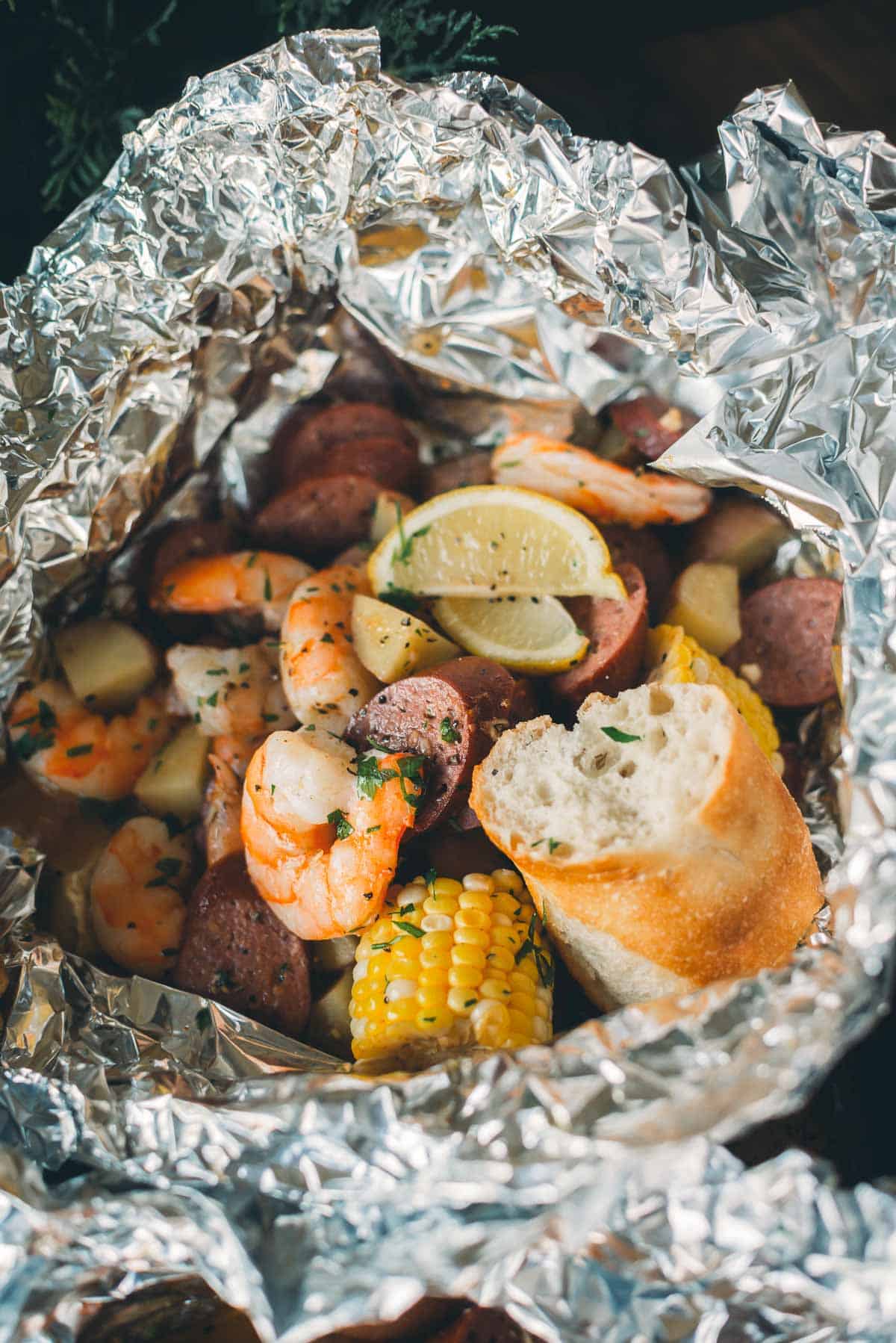 A foil packet containing cooked shrimp, sausage slices, corn on the cob, potatoes, lemon wedges, and a piece of bread.