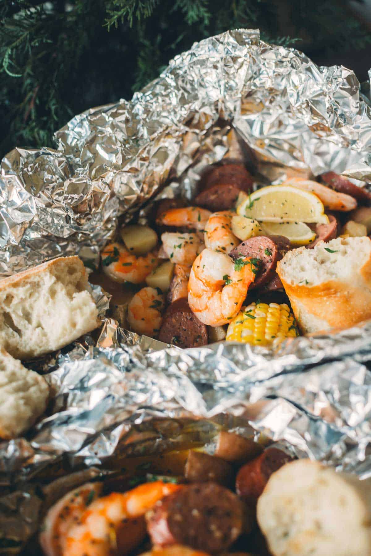 An open foil packet with shrimp, sausage, corn, potatoes, lemon slices, and bread on the side, presented on a rustic outdoor setting.