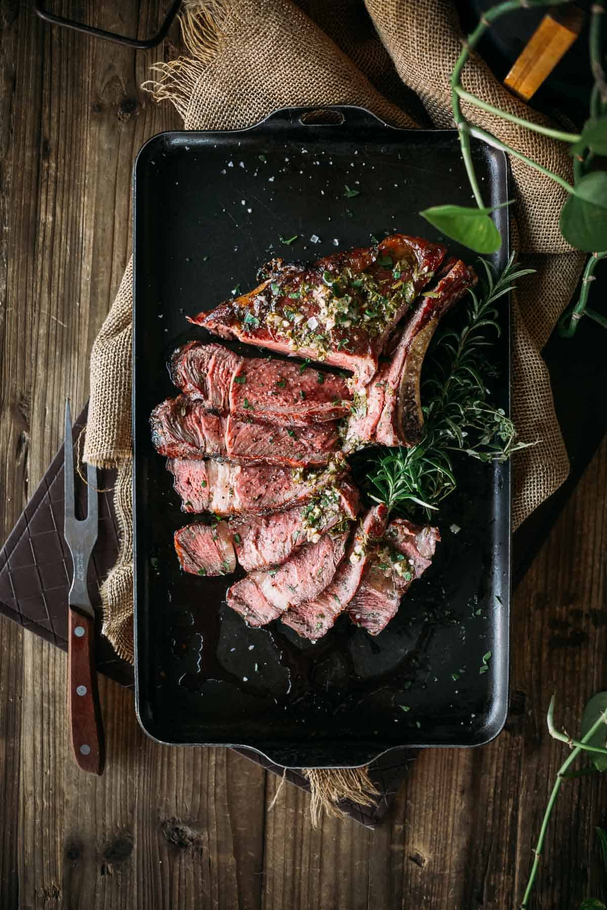 A cooked ribeye steak sliced and garnished with herbs on a black tray.