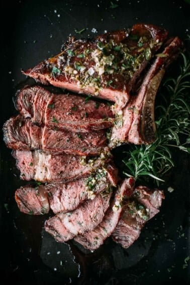 Slices of smoked steak with herbs and seasoning on a black surface, garnished with a sprig of rosemary.