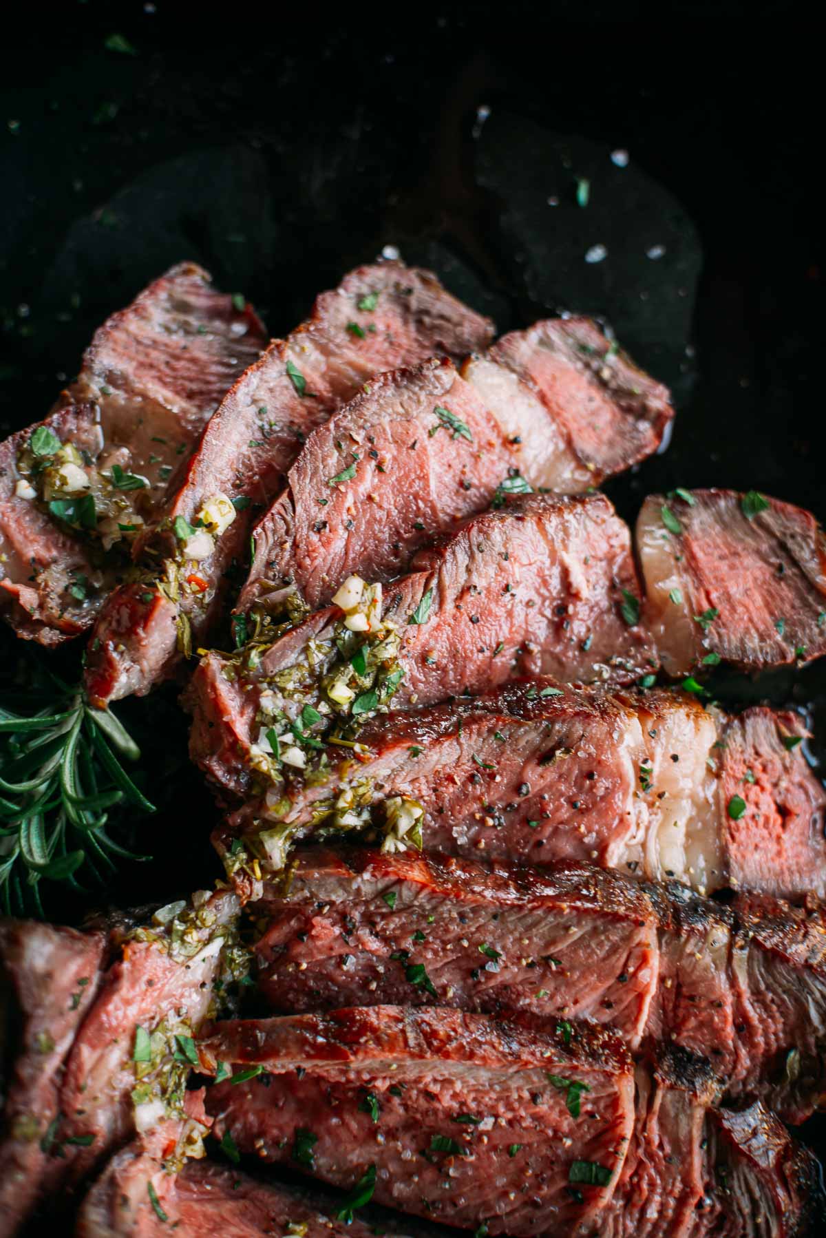 Close-up of sliced, medium-rare smoked ribeye steak garnished with herbs and garlic, displayed on a dark surface.
