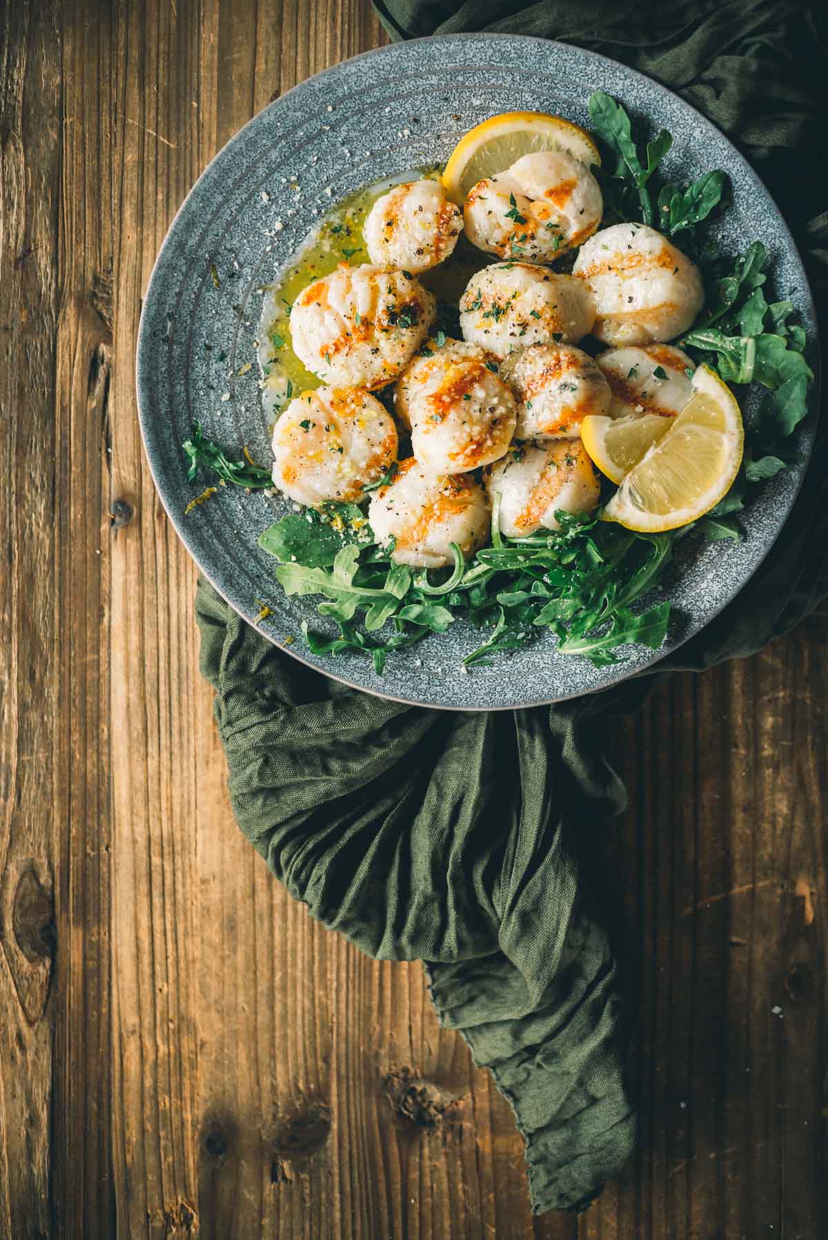 A gray plate with grilled scallops garnished with lemon wedges and greens, placed on a wooden table with a dark green cloth underneath.