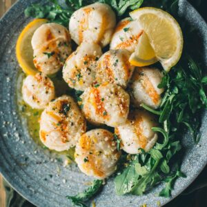 A plate of grilled scallops garnished with herbs and lemon wedges, served on a bed of arugula.