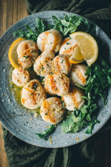 A plate of grilled scallops garnished with herbs and lemon wedges, served on a bed of arugula.