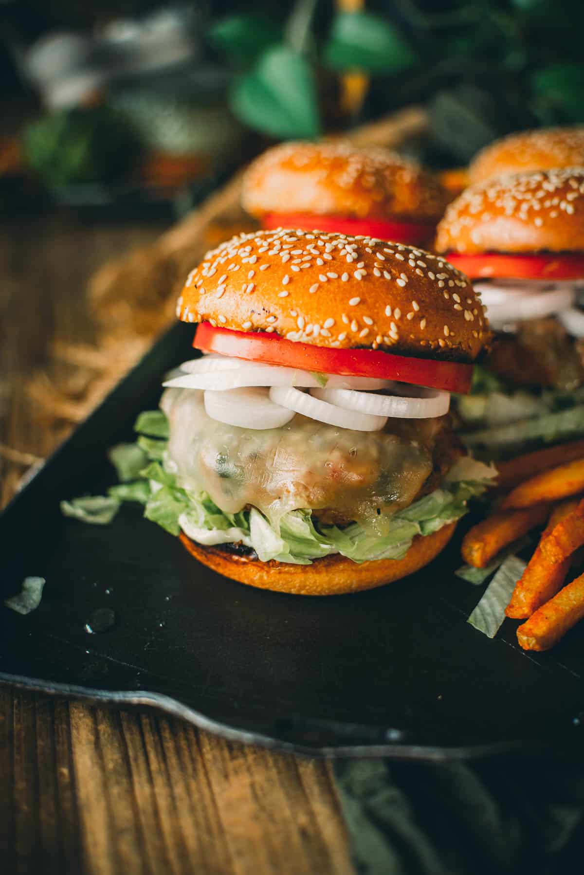 Close-up of a pork burger with lettuce, melted cheese, tomato, and onion on a sesame seed bun, served with fries on a black plate.