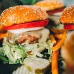 A pork burger with a sesame seed bun, lettuce, pickles, cheese, tomato, and onion slice, served with sweet potato fries on a dark plate.