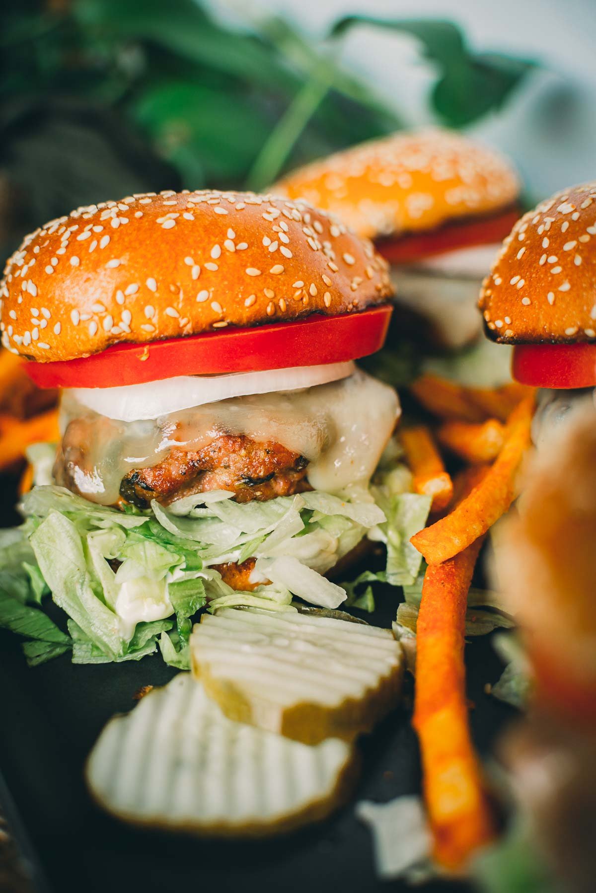 A pork burger with a sesame seed bun, lettuce, pickles, cheese, tomato, and onion slice, served with sweet potato fries on a dark plate.