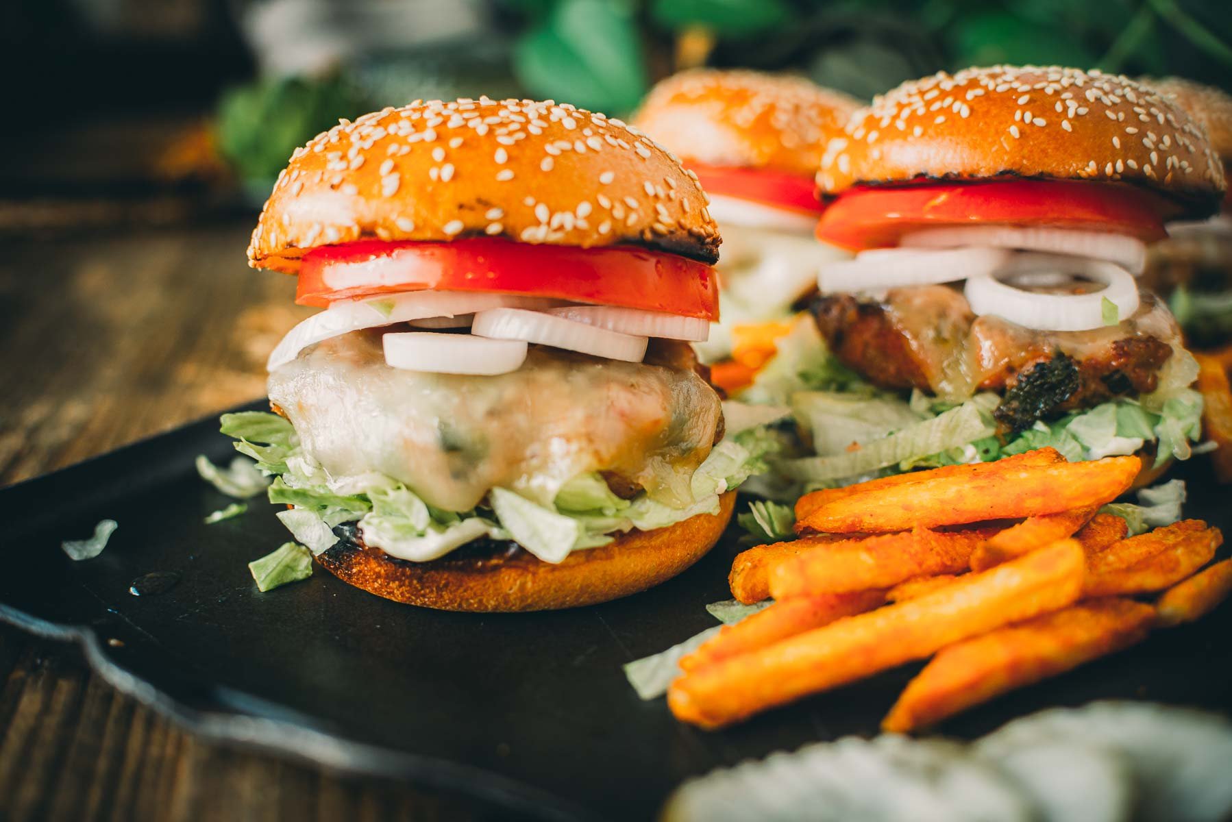 Close-up of pork burgers with lettuce, tomato, onion, and cheese, served on a black plate with a side of sweet potato fries.