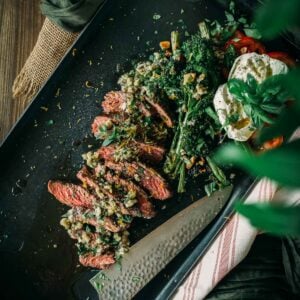 A plate with sliced steak covered in herb sauce, grilled vegetables, burrata cheese, and a knife placed nearby. The setup is on a striped cloth.