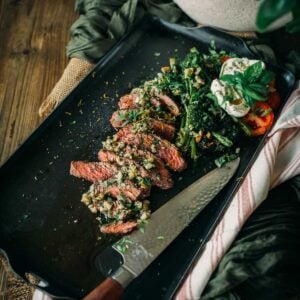 A steak dish garnished with herbs, served with a side of sautéed greens and vegetables, on a rectangular black plate beside a knife on a wooden table.