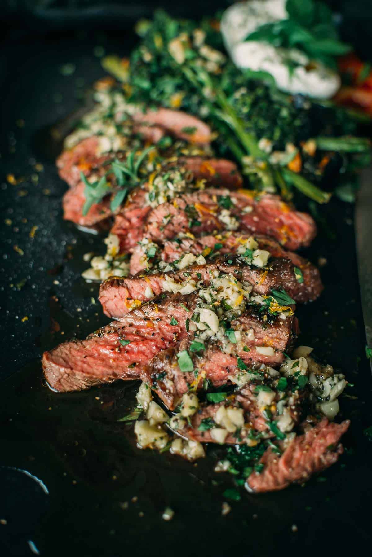 Sliced teres major steak topped with herbs and garlic, served alongside leafy greens and cheese on a dark plate.