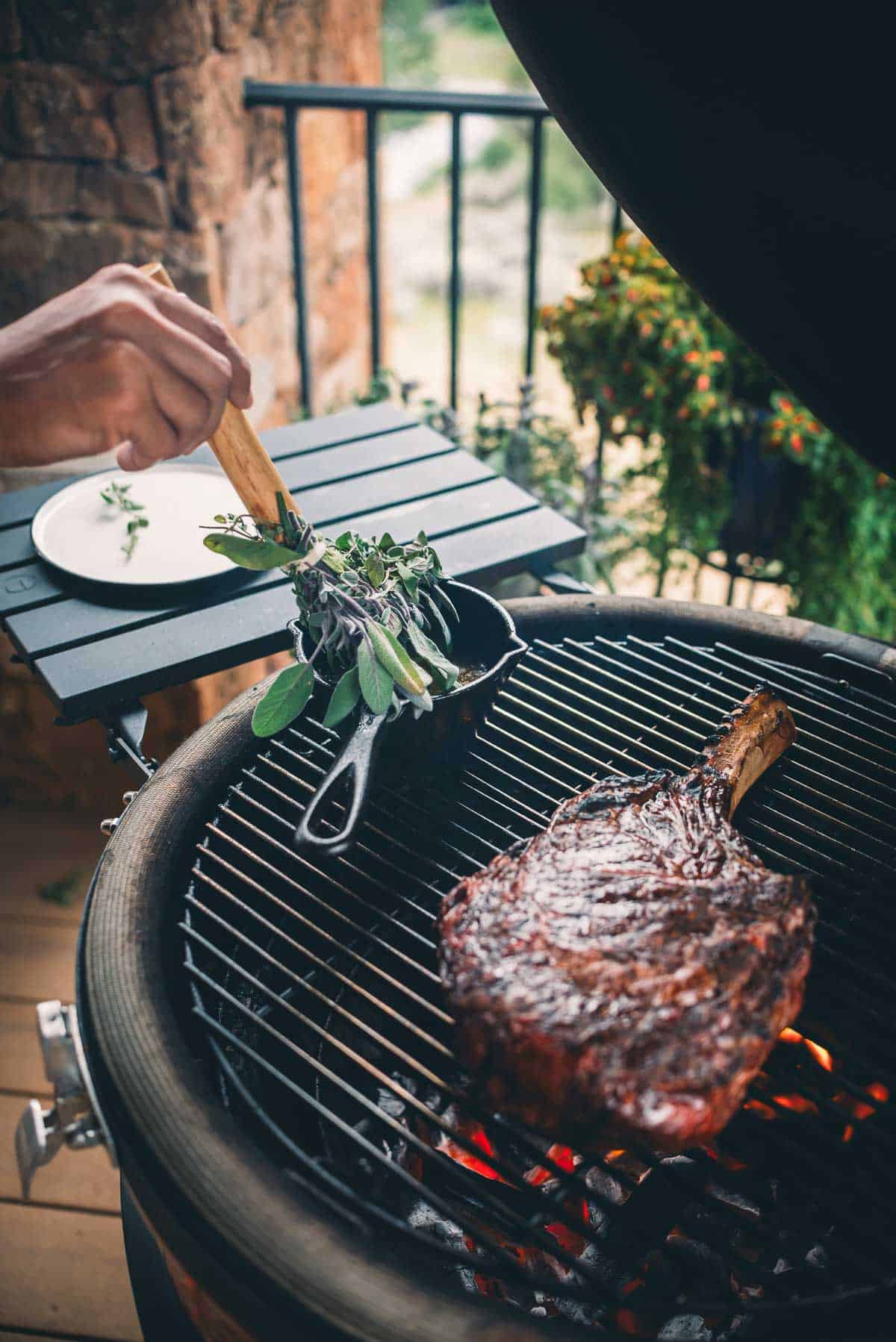 A person is basting herbs over a large piece of meat grilling on a barbecue. 