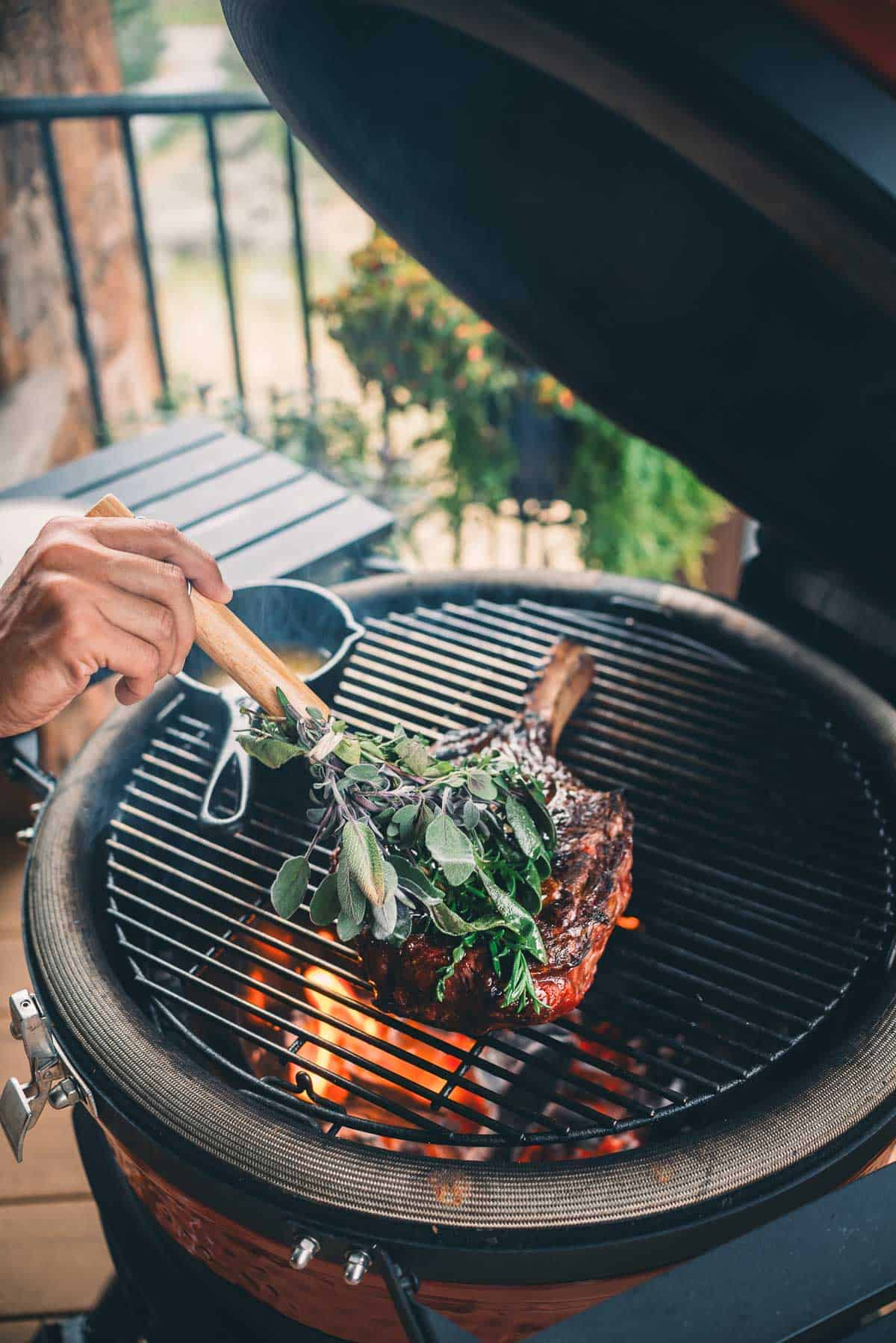 A person uses a brush made of fresh herbs to baste a large piece of meat cooking on a grill.