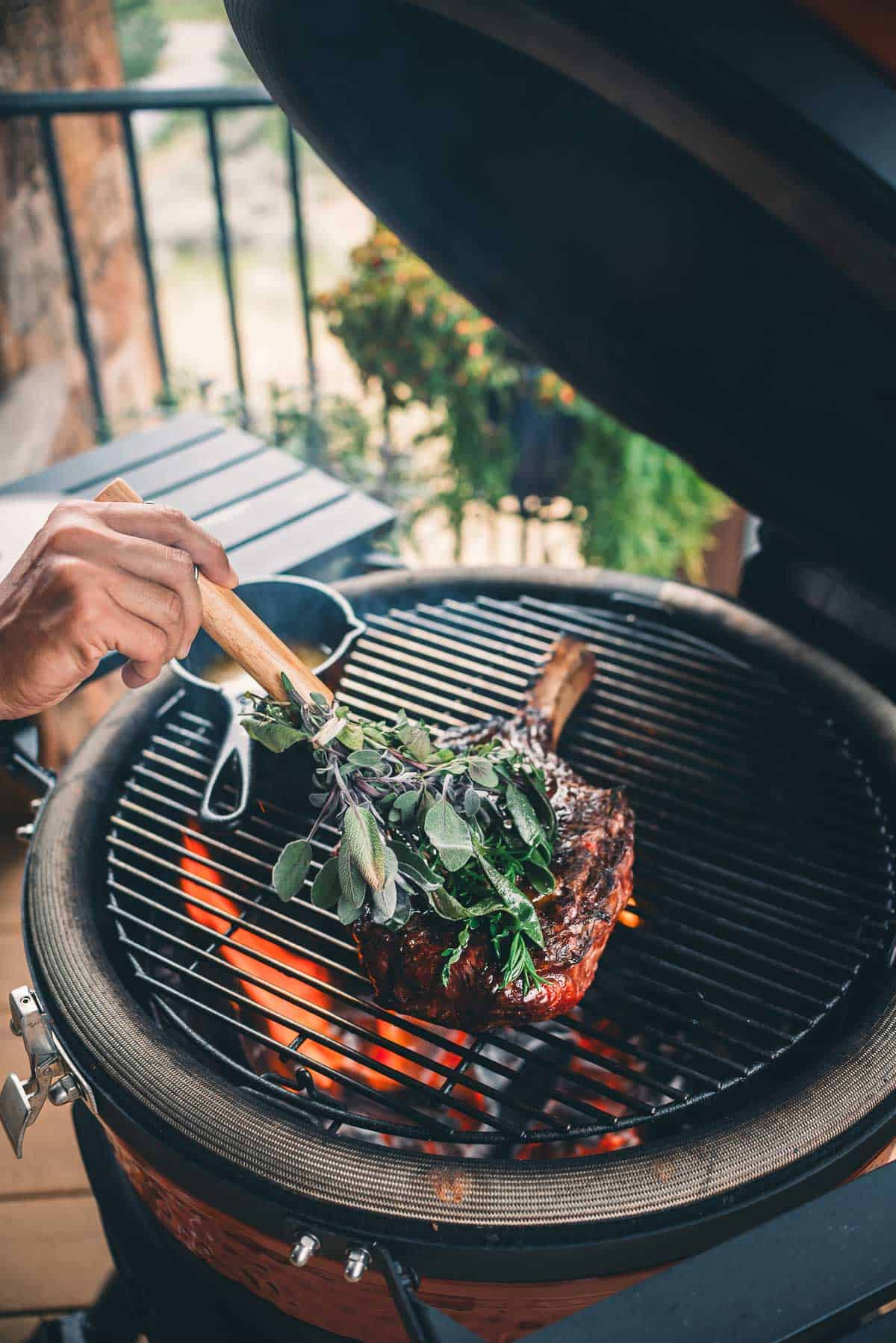 A person grills a large steak with fresh herb basting brush on a barbecue grill.