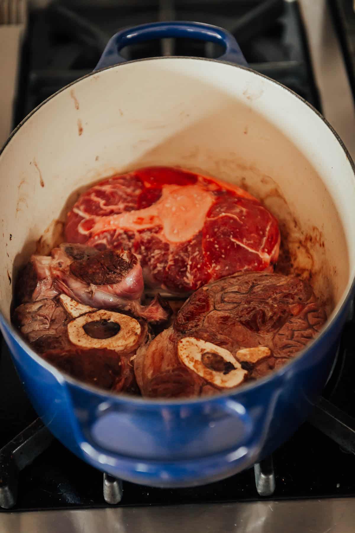 A blue pot on a stove contains three large pieces of raw meat with visible bones, ready for cooking.