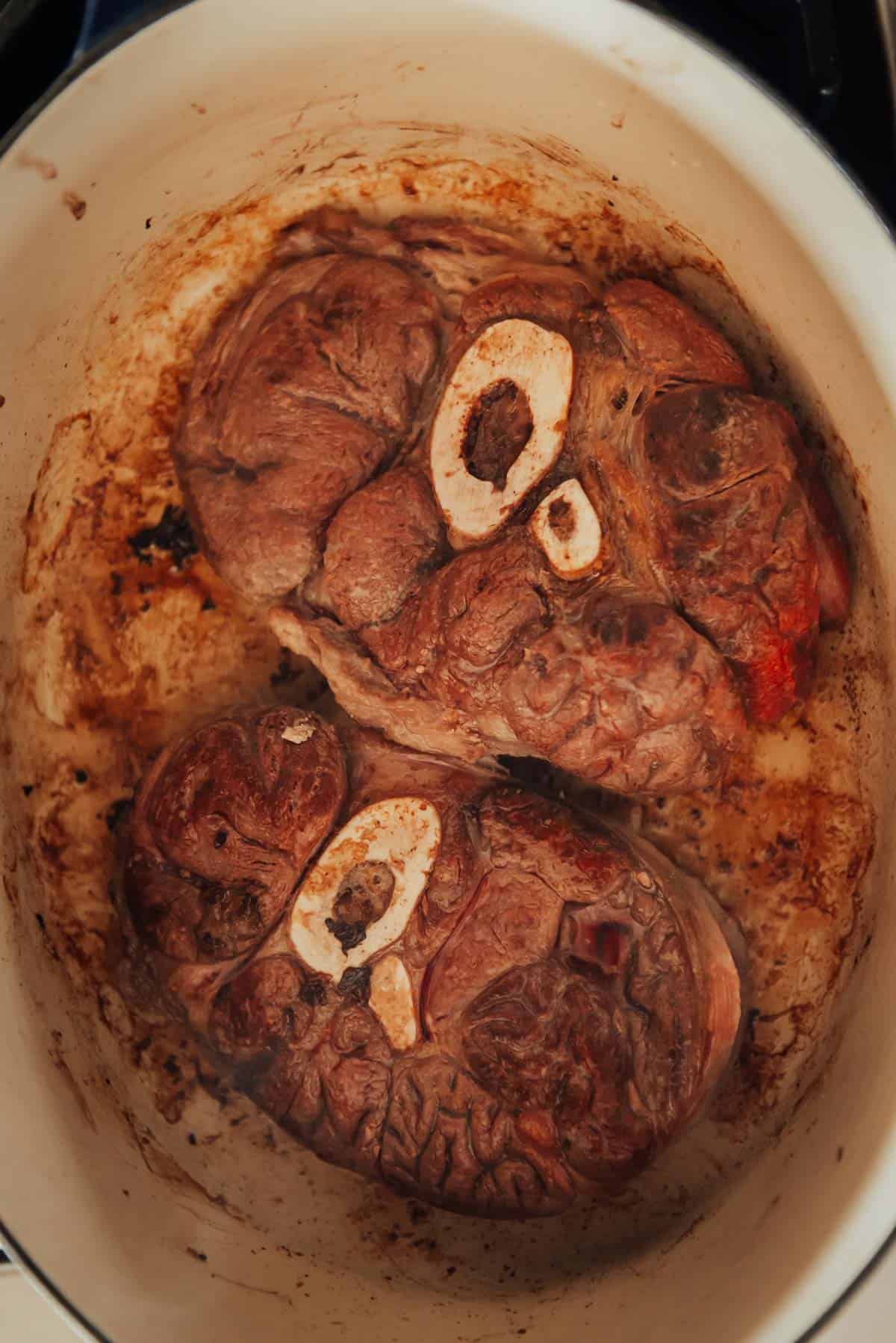 Two large pieces of meat with bone, browned and cooked, placed in a white pot. The meat is surrounded by some darkened residue from cooking.